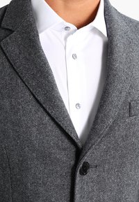 Primer plano de una persona que lleva un blazer de espiga gris sobre una camisa blanca de botones, resaltando la textura de la tela y los botones.