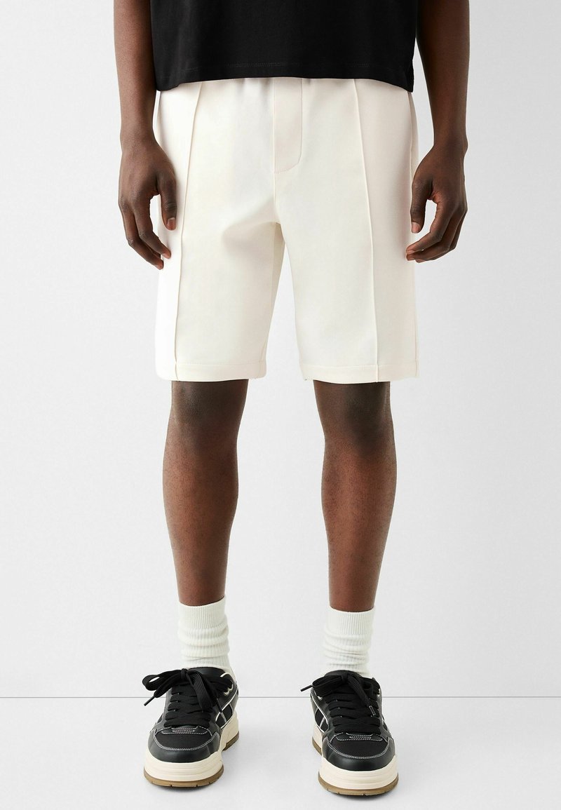 Homme portant un short blanc cintré à mi-cuisse, des chaussettes blanches à mi-mollet, des baskets noires à semelles blanches et un t-shirt noir, se tenant contre un fond blanc.