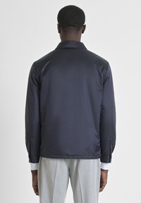 Giacca leggera navy con una texture liscia, dotata di colletto e risvolti con bottoni. Indossata su pantaloni grigi chiari.