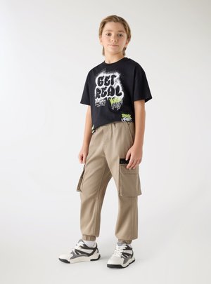 Bambino in piedi che indossa una t-shirt nera con la scritta "Get Real", pantaloni cargo beige e sneakers bianche e nere su sfondo bianco uniforme.