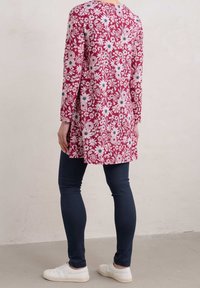 Długa bluzka w czerwonym kwiatowym wzorze z białymi i niebieskimi akcentami, wykonana z lekkiego materiału, w połączeniu z ciemnymi dopasowanymi dżinsami i białymi butami.