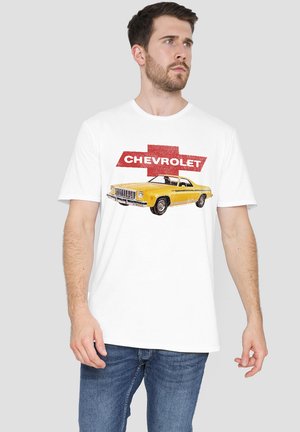 Man wearing white t-shirt with vintage yellow Chevrolet car and red Chevrolet logo, paired with blue jeans, on gray background.