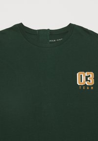 Tmavě zelené bavlněné tričko s kulatým výstřihem, na levé oblasti hrudi s bílou a oranžovou grafikou "03 1998 TEAM".