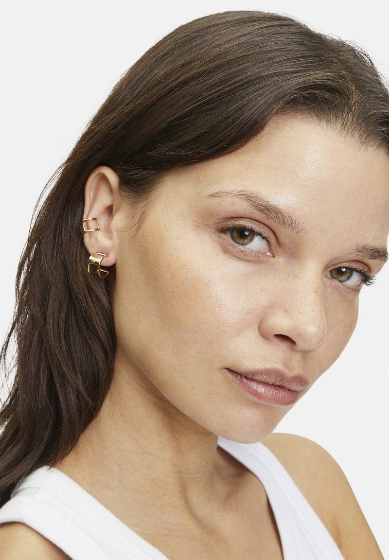 Boucles d'oreilles en métal doré avec un design géométrique comprenant deux formes carrées entrelacées. Texture lisse, apparence légère et style minimaliste.