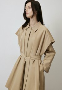 Palton bej cu brațe scurte, accesorizat cu un centură, având guler ascuțit și pliuri frontale. Textura moale și designul minimalist subliniază silueta acestuia.
