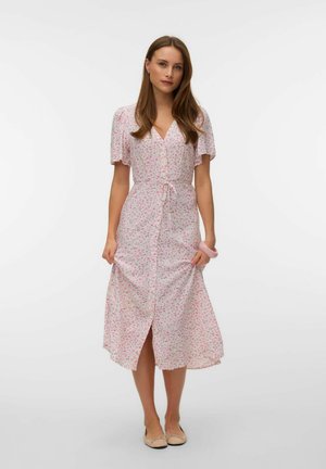 Femeie purtând o rochie midi floral roz pal, cu mâneci scurte, nasturi în față și talie cu panglică, stând în fața unui fundal alb simplu.