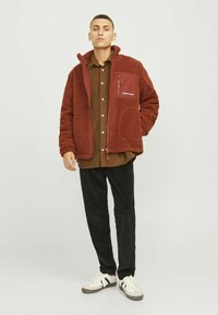 Veste en polaire rouge avec fermeture éclair à l'avant, poches latérales et poche poitrine. Portée par-dessus une chemise marron et associée à un pantalon noir et des baskets blanches.