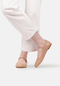 Picioare încărcate cu balerini bej cu brățări la glezne, purtate cu pantaloni albi scurtați, pe un fundal simplu.