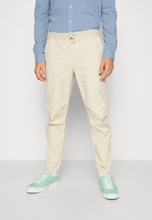 Homme portant un pantalon beige à cordon, une chemise bleu clair boutonnée et des baskets vert menthe, debout sur un fond blanc.