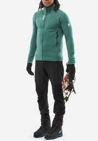 Grüne Zip-Jacke mit hohem Kragen, schwarze technische Hosen und schwarze Stiefel. Hält Kletterausrüstung und zeigt detaillierte Nähte und Texturen.