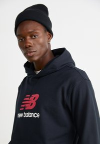 Marinefarbener Hoodie mit rotem und weißem "New Balance"-Logo auf der Vorderseite. Begleitet von einer schwarzen Strickmütze. Das Material wirkt weich und lässig.