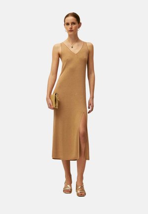 Mujer con vestido midi beige sin mangas con una abertura hasta el muslo, sosteniendo un clutch a juego y llevando sandalias doradas con tiras.