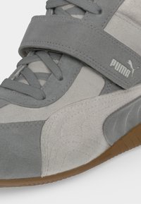 Gros plan d'une basket Puma grise et blanche avec des lacets, une bride velcro et une semelle couleur gomme.