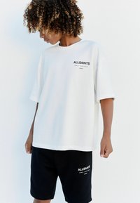 Bílý oversized tričko z bavlny s černým logem na hrudi, kombinované s černými šortkami se stejným logem blízko lemu.