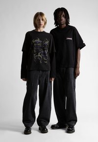 Du žmonės vilki per dideles juodas marškinėlius ir plačias juodas kelnes. Kairėje esantis marškinėlius puošia grafika, o dešinėje yra mažas logotipas. Abu dėvi juodus batus.