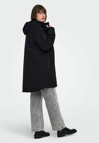 Parka negra con capucha y corte recto, que presenta un bolsillo lateral con cremallera y una tela suave; combinada con unos vaqueros de pierna ancha en gris claro y botas negras.