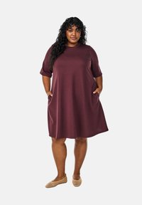 Maroon knee-length dress with short sleeves, featuring side pockets and a relaxed fit. Worn with beige flats, smooth fabric texture.