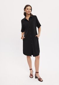 Czarna sukienka koszulowa z krótkim rękawem, z dwoma przednimi kieszeniami, luźnym fasonem i rozcięciem po boku, noszona z brązowymi sandałami na obcasie.