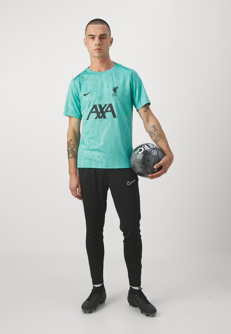 Camiseta deportiva turquesa con el logo de Liverpool, impresión negra de AXA, combinada con leggings negros y botas de fútbol negras, sosteniendo un balón de fútbol.