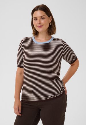 Femme aux cheveux bruns courts portant un t-shirt à manches courtes rayé marron et blanc avec un col bleu clair et un pantalon marron foncé, debout avec la main sur la hanche.