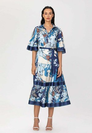 Femme debout portant une robe bleue à motifs floraux mi-longue avec col, manches larges et ceinture nouée, associée à des sandales blanches à talons.