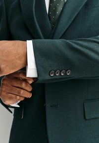 Donkergroene blazer met een zachte textuur. De witte manchet van het shirt is zichtbaar en heeft vier zwarte knopen op de mouw. Een stippenstrik is gedeeltelijk te zien.