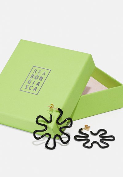 Schwarze, wellenförmige Ohrringe mit glänzender Oberfläche, neben einer limegrünen Box mit einem silbernen Logo platziert. Die Box hat ein weiches Innenleben.
