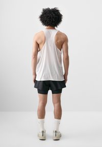 Vitt ärmlöst linne med perforerad struktur, tillsammans med svarta shorts. Modellen bär vita strumpor och beige sneakers. Bakifr�ån.
