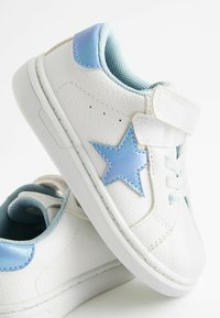 Sneakers bianchi con esterno testurizzato, dettagli blu e una stella blu. Presentano una chiusura in velcro e foderatura in tessuto.