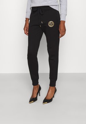 Kobieta ubrana w czarne wąskie spodnie dresowe z złotą naszywką na udzie oraz czarne szpilki ze złotymi końcówkami, w połączeniu z jasnoszarym topem.