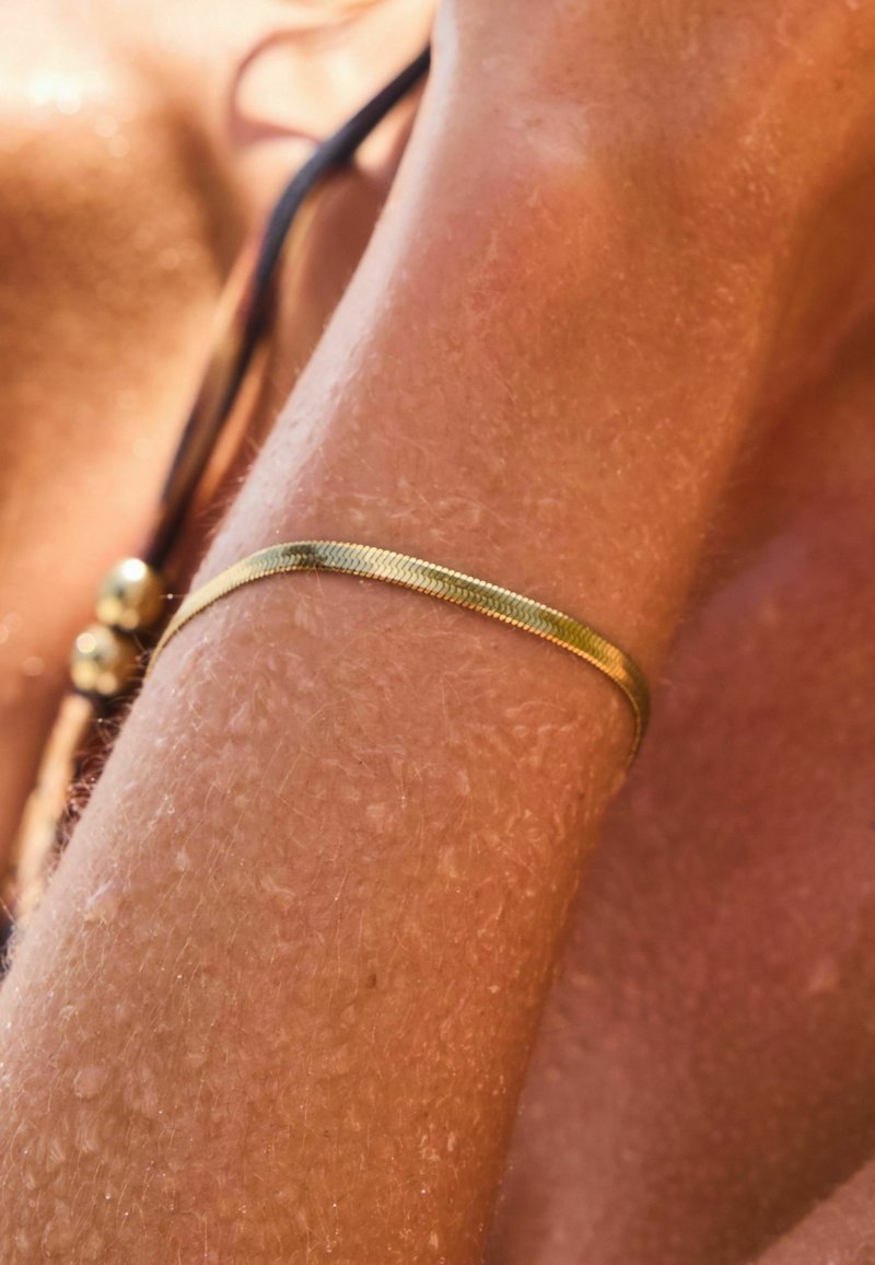 Bracciale color oro con una superficie testurizzata, caratterizzato da un design slim e piatto che si avvolge attorno al polso, posizionato su una pelle abbronzata.