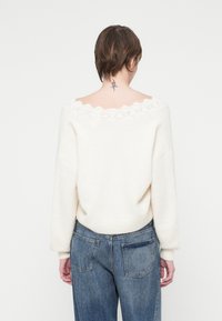 Weicher, cremefarbener Pullover mit einem gewellten Spitzenhalsausschnitt, weiten Ärmeln und einer lockeren Passform, kombiniert mit hochsitzenden blauen Jeans.