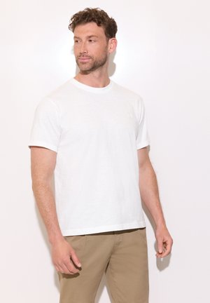Mand med kort krøllet hår og skæg iført en ensfarvet hvid kortærmet t-shirt og beige bukser, kigger til venstre mod en ensfarvet baggrund.