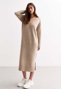 Beige gebreide jurk met V-hals, geribbelde mouwen, zijsplitten en een lengte tot halverwege de kuit, gecombineerd met witte sneakers op een neutrale achtergrond.