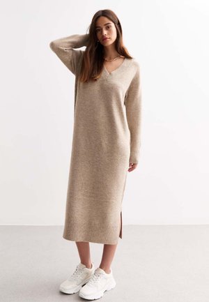 Beige gebreide jurk met V-hals, geribbelde mouwen, zijsplitten en een lengte tot halverwege de kuit, gecombineerd met witte sneakers op een neutrale achtergrond.
