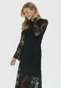 Juoda nėriniuota suknelė su ilgomis permatomomis rankovėmis, gėlių raštu ir prigludusia krūtine. Sijonas yra pusiau skaidrus, suteikiantis elegancijos.