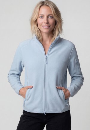 Blonde Frau trägt eine hellblaue Fleecejacke mit Reißverschluss und hat die Hände in den Taschen, sie steht vor einem einfarbigen hellgrauen Hintergrund.