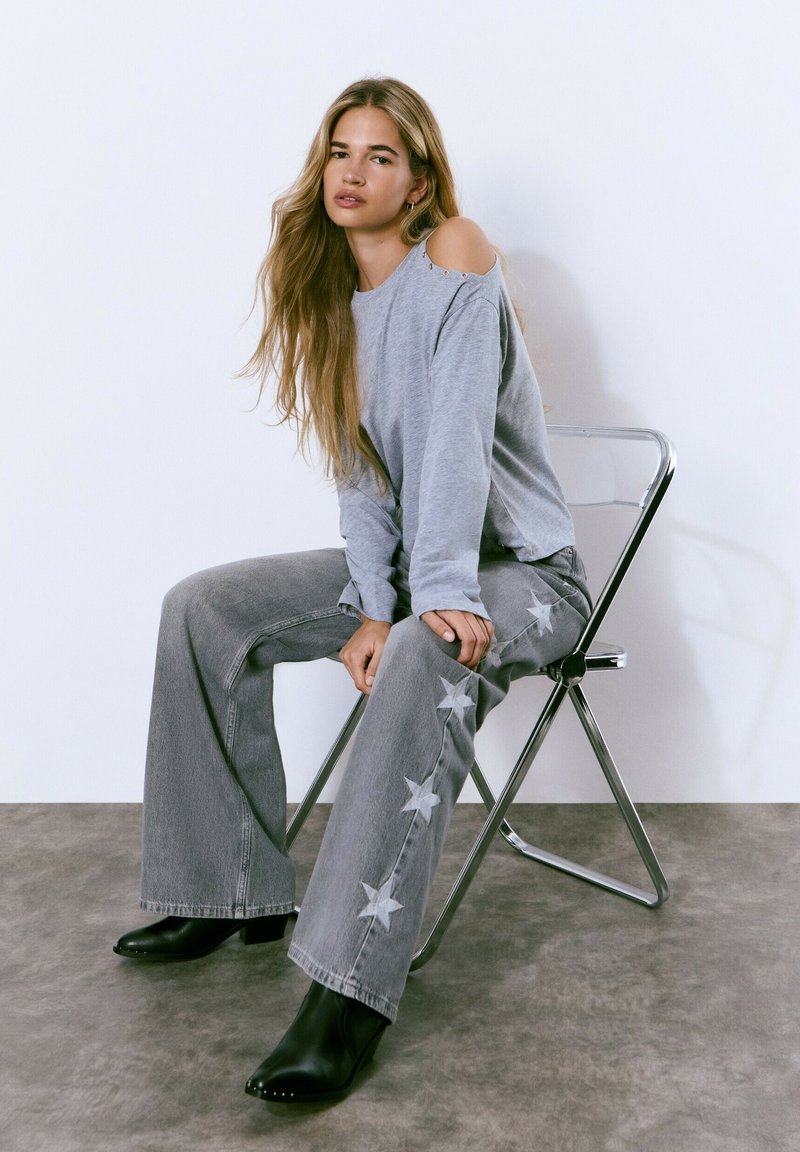 Camisa gris de manga larga con corte en los hombros, combinada con jeans grises de pierna ancha con apliques de estrellas. Botines negros de punta. Silla plateada.