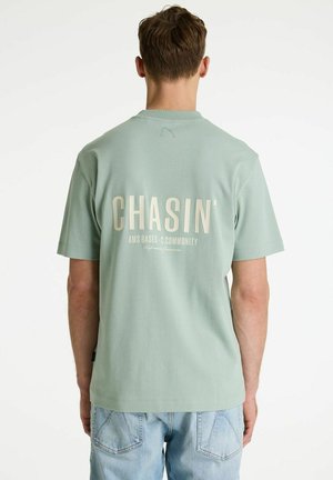 Moški s kratkimi rjavimi lasmi, oblečen v mintasto zeleno majico s tekstom "CHASIN'" in svetlo modre kavbojke, stoji s hrbtom obrnjeno proti beli podlagi.