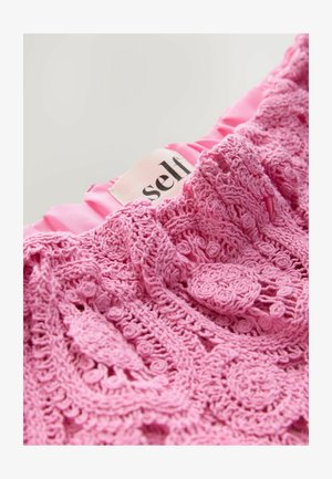 Tissu au crochet rose avec des motifs floraux complexes et une bordure ondulée lisse et brillante de couleur rose. Présente une étiquette indiquant "auto."
