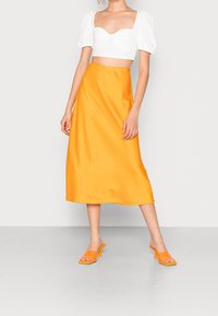 Bílý crop top s balónovými rukávy spárovaný s jasně oranžovou saténovou midi sukňí a odpovídajícími muly na blokovém podpatku, které ukazují hladké textury.