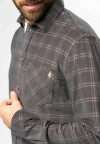 Camisa de manga larga a cuadros grises hecha de una tela suave, con un bolsillo en el pecho. Cuenta con acentos de color marrón claro y un pequeño detalle bordado.