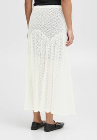 Jupe maxi en dentelle de couleur blanc cassé ; dispose d'une taille ajustée et d'un design fluide avec un motif floral et des franges. Chaussures plates noires portées.