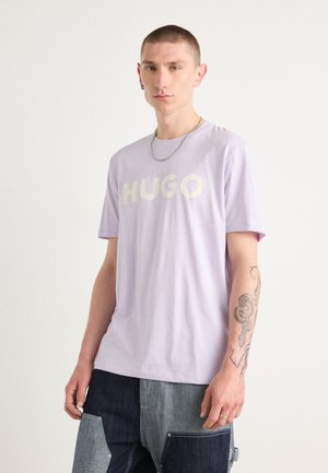 Lys lavendelfarvet t-shirt med et stort hvidt "HUGO" logo på fronten, korte ærmer og en glat bomuldsstruktur.