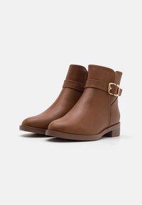 Bottes chelsea en cuir brun avec un bout arrondi, un talon empilé et une bouclerie décorative sur le côté. Texture lisse et design minimaliste.