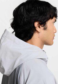 Prenda con capucha de color gris claro con una textura suave, que presenta un cordón en el cuello y una capucha curva. Vista de perfil en primer plano con cabello oscuro y desestructurado.