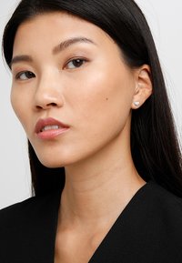 Sölvfärgade hjärtformade örhängen med texturerad design, bärs av en person med långt, mörkt hår, mot en enfärgad bakgrund. Mörka kläder syns.