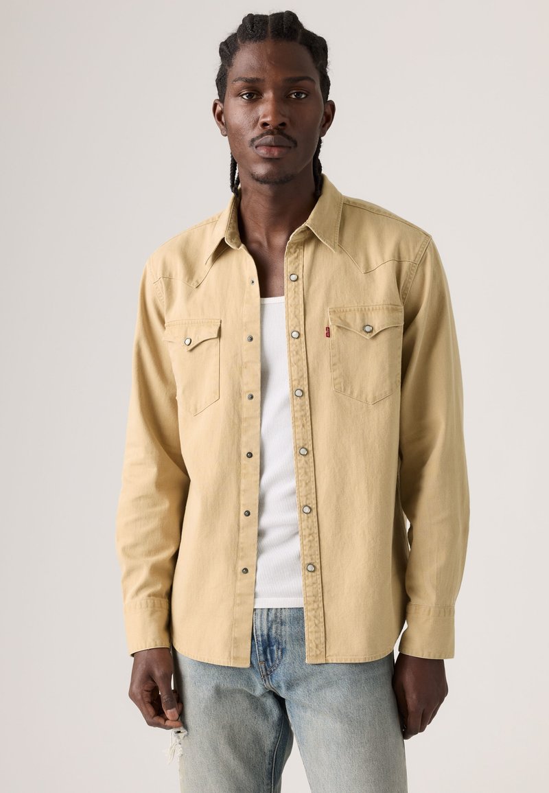 Homme aux cheveux tressés portant une chemise beige déboutonnée sur un débardeur blanc et un jean bleu clair déchiré, debout devant un fond uni.