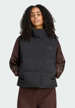 Kvinde med krøllet hår iført en sort polstret vest over en brun bluse med lange ærmer med mønster mod en ensfarvet baggrund.