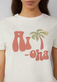 Roomkleurige katoen t-shirt met een speelse grafische druk van de tekst "Aloha" in koraal en groen, aangevuld met een ontwerp van een palmboom en zon.